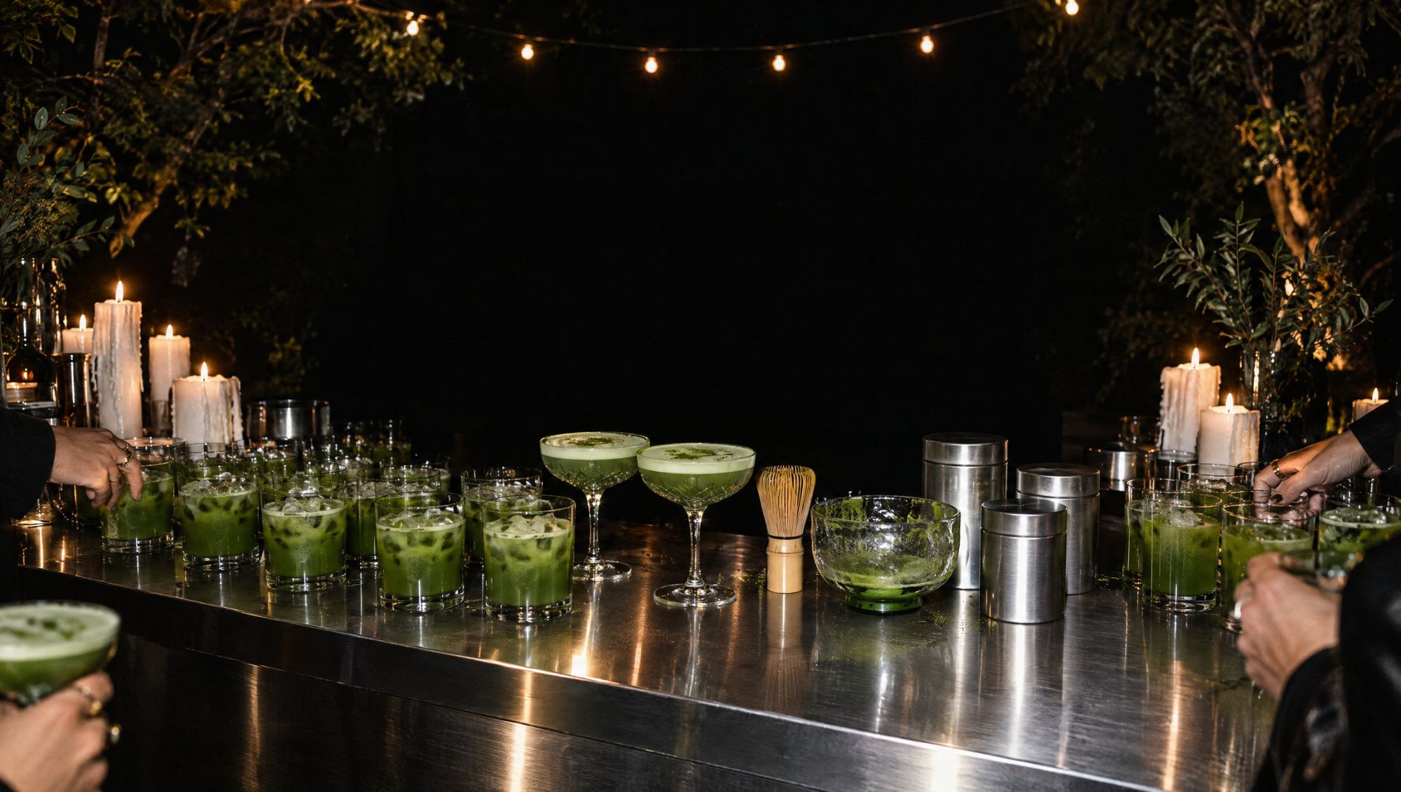 A long steel bar at night lined with iced matcha, crystal coupes and candles, with guests reaching in